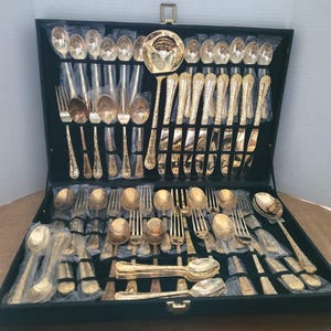 May include: A complete gold-colored flatware set displayed in a black case. The set includes spoons, forks, knives, and serving utensils. The utensils have decorative handles and are arranged neatly within the case.