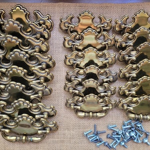 May include: A collection of ornate brass drawer pulls with a decorative, scrolled design. The pulls are arranged in stacks and rows, with a pile of silver screws at the bottom. The hardware is set against a neutral, textured background.