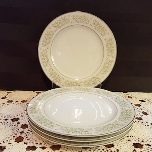 Puede incluir: Juego de cuatro platos de cena de cerámica blanca con un patrón floral verde alrededor del borde. Los platos tienen un borde dorado.