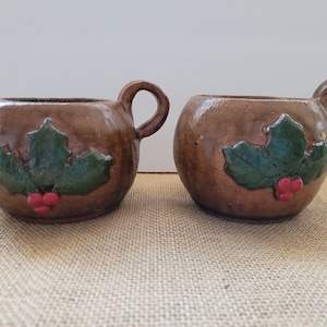 May include: Two brown ceramic mugs with green holly leaf and red berry designs.