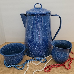 Peut inclure: Une cafetière en émail bleu moucheté avec un couvercle, un petit pichet et une tasse. L'ensemble est de style vintage. La cafetière mesure environ 25 cm de haut. L'ensemble est sur une surface en toile de jute avec des perles rouges, blanches et bleues.