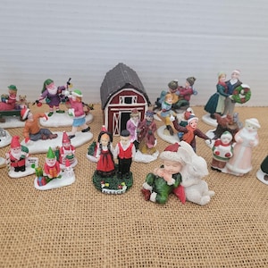 Op de afbeelding: Een verzameling miniatuur kerstdorpfiguren. De beeldjes beelden verschillende winterse taferelen uit, waaronder mensen die schaatsen, kerstbomen dragen en een rode schuur. De figuren zijn geschilderd in heldere kleuren en staan op witte voetstukken.