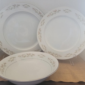 May include: A set of three white porcelain dishes. Two dinner plates and one bowl are displayed. Each piece has a gold floral pattern around the rim and a thin gold band. The dishes are on clear plastic stands.