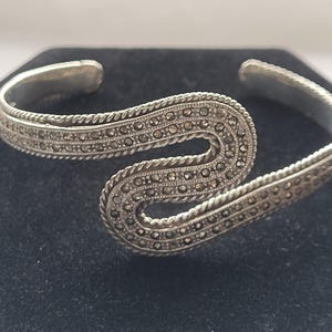 May include: A silver-toned cuff bracelet with a wave-like design. The bracelet is adorned with small, dark-colored stones and a twisted rope-like border. The open-ended design allows for easy wear.