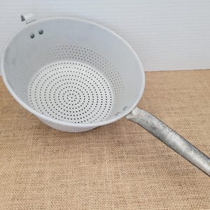 May include: A light gray metal colander with a long, weathered silver-toned handle. The bowl features a perforated base with numerous small circular holes for drainage and a small metal loop at the top edge for hanging storage purposes.