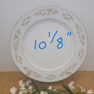 May include: A white ceramic plate with a floral design and a silver rim. The plate measures 25.7 cm in diameter. The floral pattern features beige and white flowers. The plate is displayed on a clear stand.