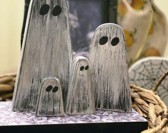 May include: A set of four wooden ghost figures with a distressed white and black finish. Each ghost has two black oval eyes. The ghosts are arranged on a decorative box with a floral pattern, inside a woven basket, perfect for Halloween.