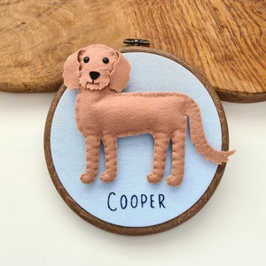 May include: A handmade felt dog in a wooden embroidery hoop. The light brown dog is stitched onto a light blue fabric, with the name "COOPER" embroidered below. The hoop is a dark brown color.