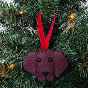 May include: A brown felt dog ornament with black eyes and a black nose. The ornament is hanging from a red ribbon on a green Christmas tree.