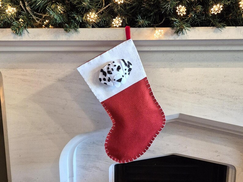 May include: A red and white Christmas stocking with a felt dalmatian dog face on the front. The stocking is hanging on a white mantel.