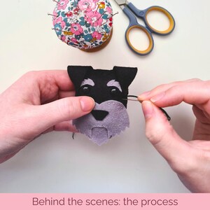 May include: A close-up of a hand sewing black thread onto a felt dog face. The dog has black fur, a purple beard, and black eyes. The hand is holding a needle and thread.