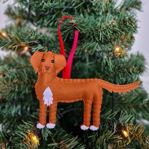 May include: A felt Christmas bauble shaped like a brown dog with white markings. The bauble is hanging from a red ribbon on a green Christmas tree.