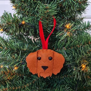 May include: A felt Christmas ornament shaped like a brown dog's head with black eyes and a black nose. The ornament is hanging from a red ribbon on a green Christmas tree.