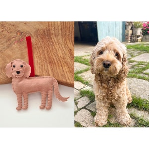 May include: A felt dog ornament in a light brown color with black eyes and a red ribbon. The ornament is on a wooden surface. A real dog of the same breed is sitting on a stone path.
