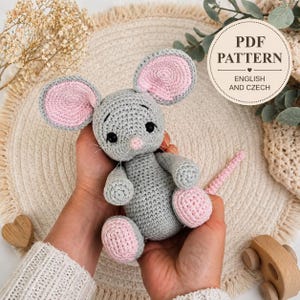 May include: A gray crocheted mouse toy with large pink inner ears, a small pink nose, and black safety eyes. The toy features a long pink tail and a cylindrical body. A circular label in the upper right corner reads PDF PATTERN ENGLISH AND CZECH.