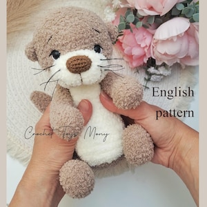 May include: A handmade crochet otter toy in shades of brown and white. The otter has black eyes, a brown nose, and long whiskers. The text "English pattern" is visible. The toy is held in someone's hands.