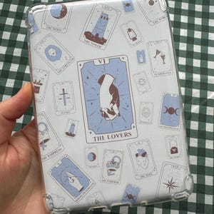 May include: A clear phone case with a white background and a repeating pattern of tarot cards. The cards are illustrated with various symbols and text, including "The Lovers", "The Tower", "The Empress", "The Fool", and "The World".