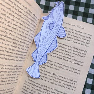 May include: A blue and white fish-shaped bookmark. The bookmark is a detailed illustration of a fish, with fins, scales, and a tail. The bookmark is placed on an open book with text.