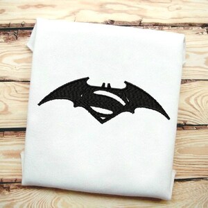 May include: Black embroidered Superman logo with a bat wing design on a white t-shirt.