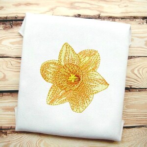 Può includere: Un singolo fiore di narciso giallo ricamato su uno sfondo di tessuto bianco.