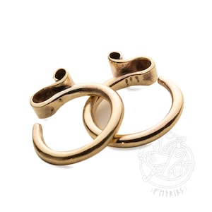 Op de afbeelding: Twee gouden metalen ringen met een uniek, gebogen ontwerp. De ringen zijn in elkaar gehaakt en lijken gemaakt te zijn van glad, gepolijst metaal.
