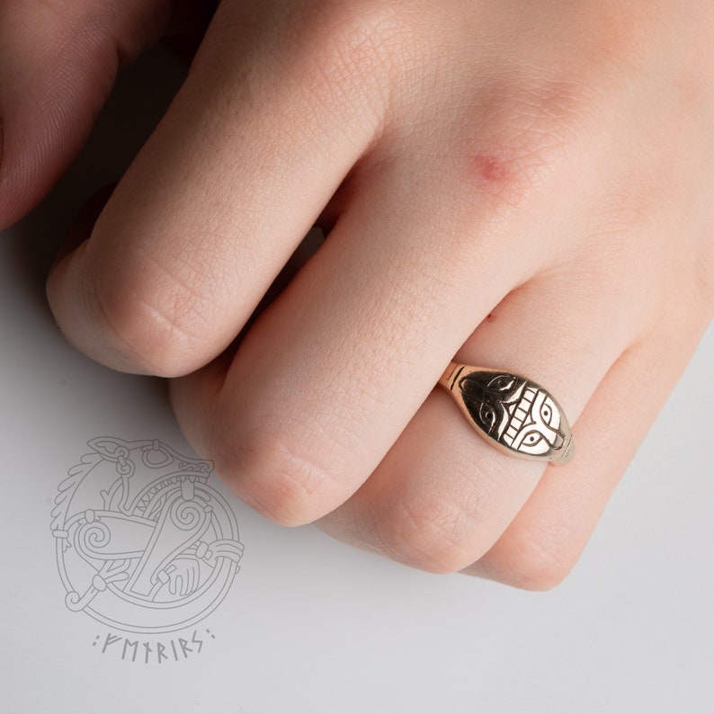 Op de afbeelding: Een gouden ring met een gedetailleerd ontwerp van een gezicht op de bovenkant. De ring wordt gedragen aan een vinger.