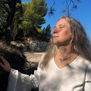 K&ouml;nnte beinhalten: Eine Frau mit langem grauem Haar tr&auml;gt ein wei&szlig;es Hemd mit einer goldenen Halskette mit drei Runensymbolen. Die Halskette tr&auml;gt den Text "Norse Runes Wisdom by Fenrir's". Die Frau steht in einem Wald mit B&auml;umen und blauem Himmel im Hintergrund.