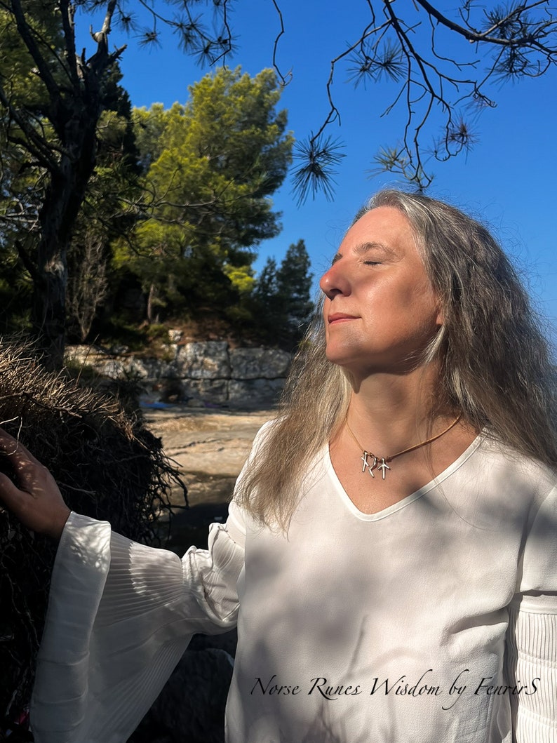 K&ouml;nnte beinhalten: Eine Frau mit langem grauem Haar tr&auml;gt ein wei&szlig;es Hemd mit einer goldenen Halskette mit drei Runensymbolen. Die Halskette tr&auml;gt den Text "Norse Runes Wisdom by Fenrir's". Die Frau steht in einem Wald mit B&auml;umen und blauem Himmel im Hintergrund.