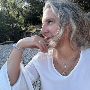 K&ouml;nnte beinhalten: Eine Frau mit langen, grauen Haaren tr&auml;gt ein wei&szlig;es Hemd mit einer silbernen Halskette mit einem Runen-Anh&auml;nger. Die Halskette ist mit einer zweiten, d&uuml;nneren Kette &uuml;bereinander gelegt. Die Frau sitzt auf einem Bett aus kleinen, grauen Steinen. Der Text "Norse Runes Wisdom by FenrirS" ist am unteren Rand des Bildes sichtbar.