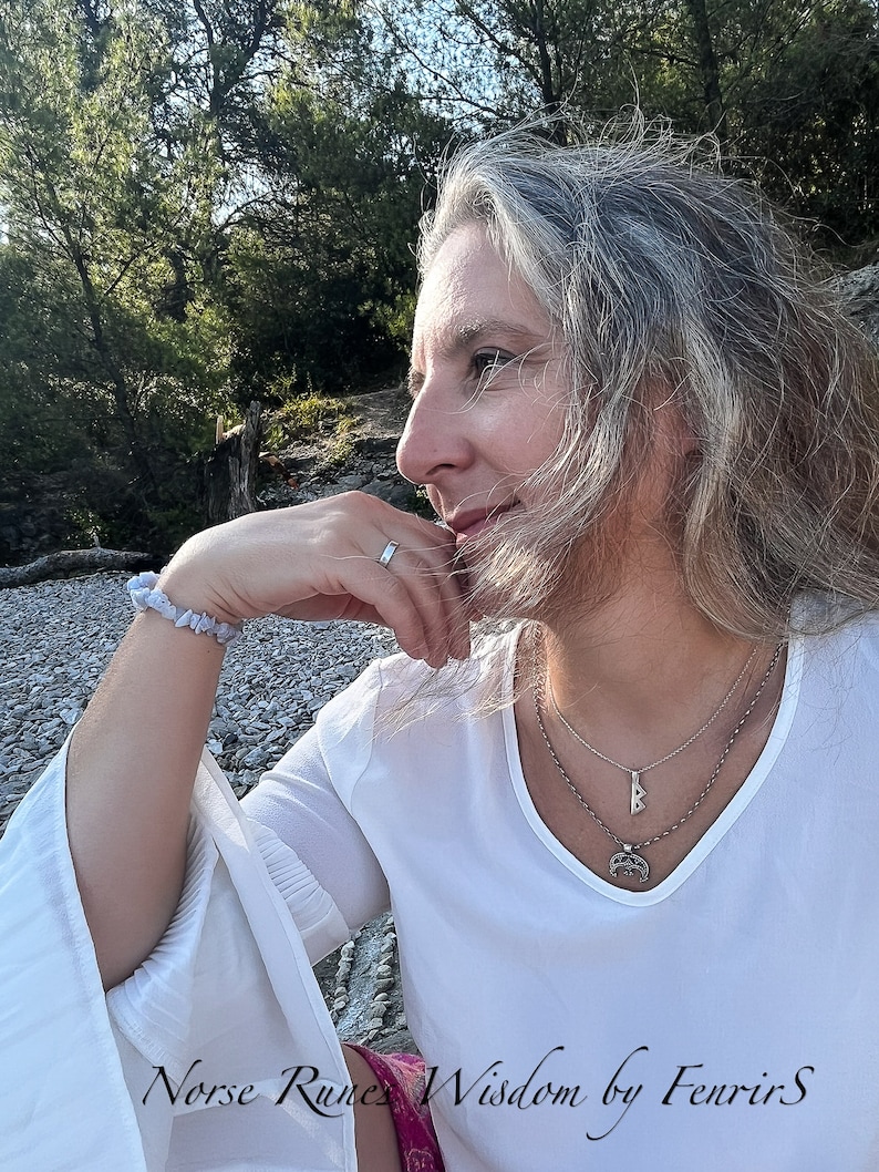 K&ouml;nnte beinhalten: Eine Frau mit langen, grauen Haaren tr&auml;gt ein wei&szlig;es Hemd mit einer silbernen Halskette mit einem Runen-Anh&auml;nger. Die Halskette ist mit einer zweiten, d&uuml;nneren Kette &uuml;bereinander gelegt. Die Frau sitzt auf einem Bett aus kleinen, grauen Steinen. Der Text "Norse Runes Wisdom by FenrirS" ist am unteren Rand des Bildes sichtbar.