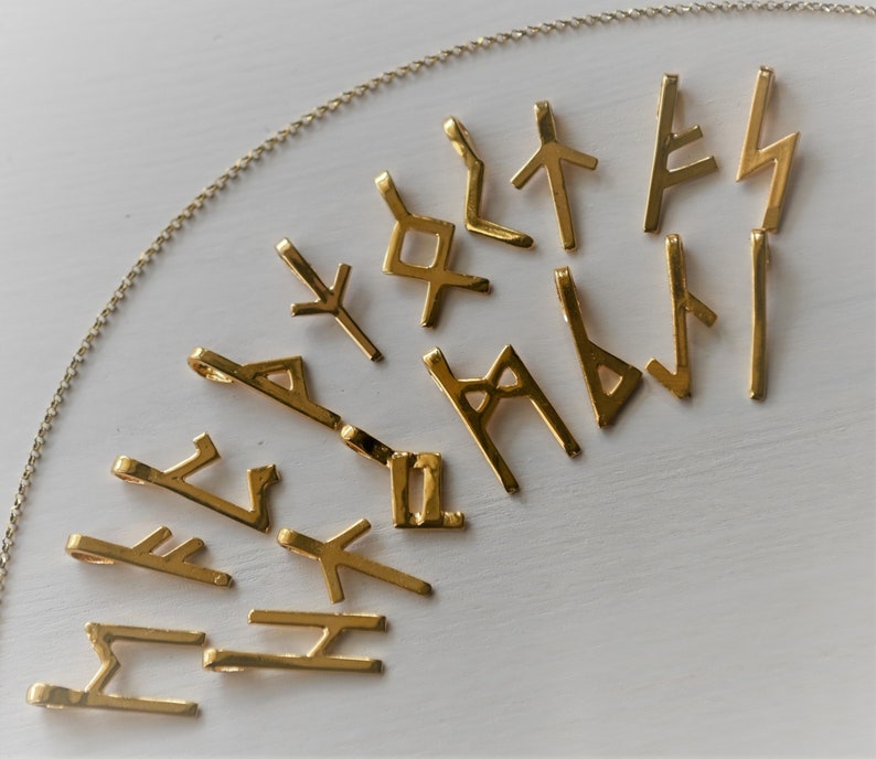 K&ouml;nnte beinhalten: Eine Sammlung goldfarbener Metallanh&auml;nger in Form von Runen, einer Art alter Schrift. Die Anh&auml;nger sind in einem Halbkreis auf einer wei&szlig;en Oberfl&auml;che angeordnet.