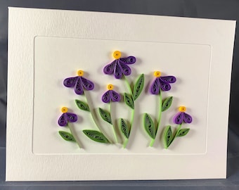 May include: A white card with a purple and green paper flower arrangement. The flowers have yellow centers and are arranged in a cluster on the card.