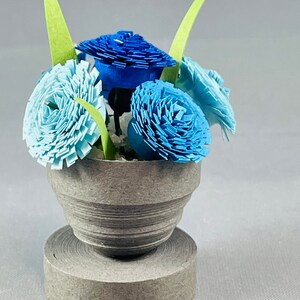 May include: A small gray paper mache planter with a blue and light blue paper flower arrangement. The flowers are made of paper and are arranged in a bouquet.