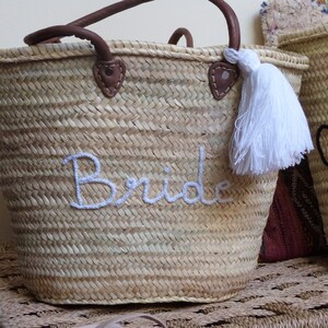May include: A woven straw tote bag with leather handles and a white tassel. The bag is embroidered with the word "Bride" in white thread.