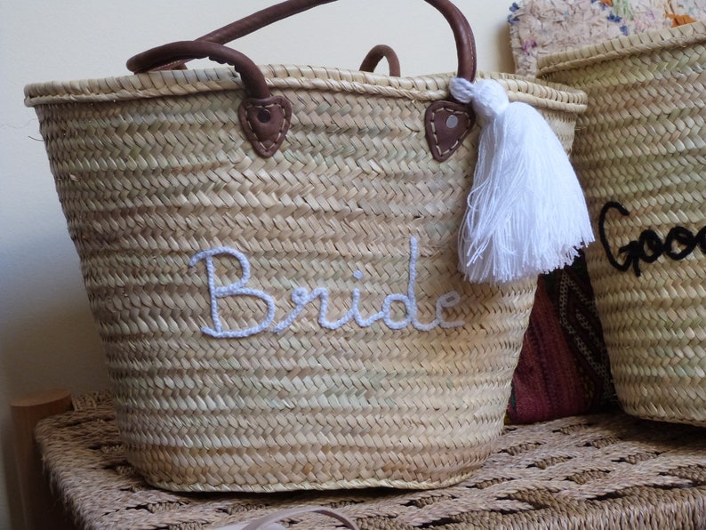 May include: A woven straw tote bag with leather handles and a white tassel. The bag is embroidered with the word "Bride" in white thread.