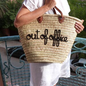 May include: A straw tote bag with a black leather handle and the words "out of office" embroidered in black thread.