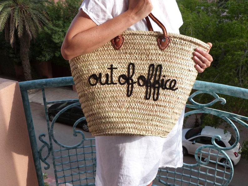 May include: A straw tote bag with a black leather handle and the words "out of office" embroidered in black thread.