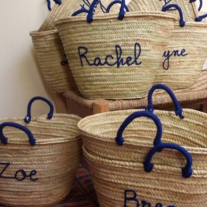May include: Five straw tote bags with blue handles and personalized names. The names are "Zoe", "Brea", "Rachel", "yne", and a partially visible name.