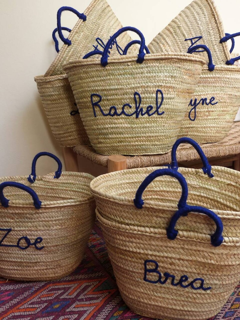 May include: Five straw tote bags with blue handles and personalized names. The names are "Zoe", "Brea", "Rachel", "yne", and a partially visible name.