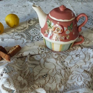 May include: A pink and white teapot with floral designs sits on a white lace tablecloth with two lemons and biscotti beside it.