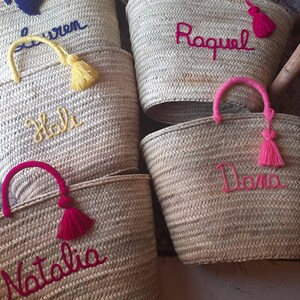 May include: Five straw tote bags with personalized names embroidered in colorful thread. The bags have different colored tassels and handles. The names are "Holly", "Lauren", "Raquel", "Dana", and "Natalia".