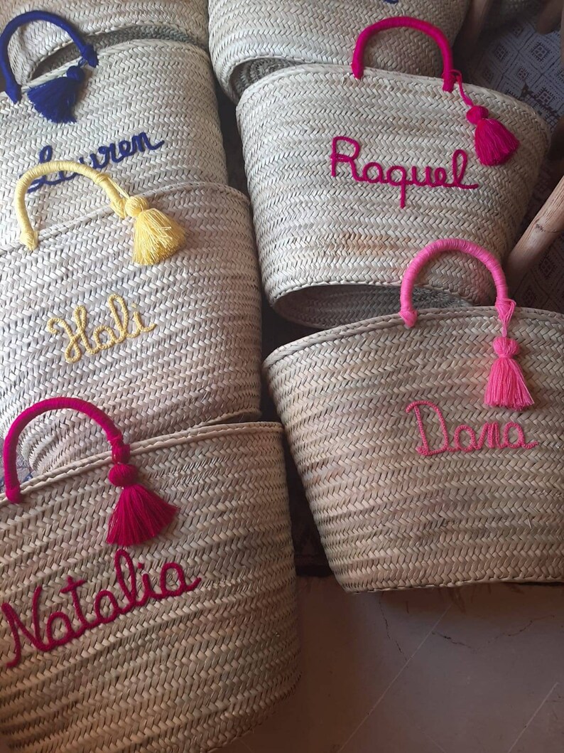 May include: Five straw tote bags with personalized names embroidered in colorful thread. The bags have different colored tassels and handles. The names are "Holly", "Lauren", "Raquel", "Dana", and "Natalia".
