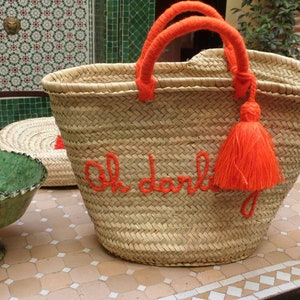 May include: A large, woven straw tote bag with orange handles and a tassel. The bag has the phrase "Oh darling" embroidered on the front in orange thread.
