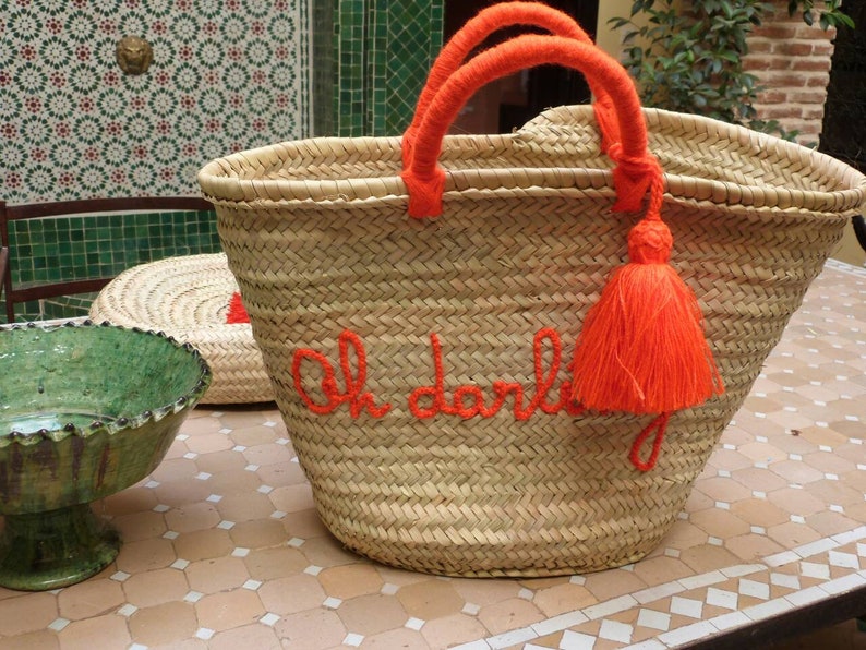 May include: A large, woven straw tote bag with orange handles and a tassel. The bag has the phrase "Oh darling" embroidered on the front in orange thread.