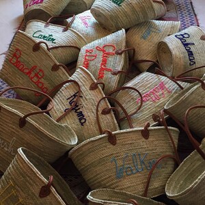 May include: A collection of straw tote bags with various embroidered names, including "Corbin", "Glenda", "Harvill", "Black Bag", "Kitima", "Walker", "Lisle", and "Balomas".