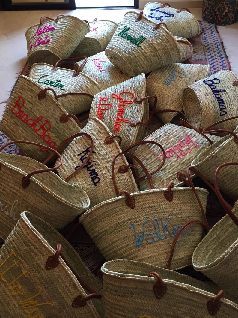 May include: A collection of straw tote bags with various embroidered names, including "Corbin", "Glenda", "Harvill", "Black Bag", "Kitima", "Walker", "Lisle", and "Balomas".