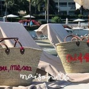 May include: Two large straw tote bags with leather handles. One bag has the text "you, me, sea" embroidered in pink thread. The other bag has the text "Oui, oui, oui" embroidered in pink thread. Both bags are sitting on white beach chairs.
