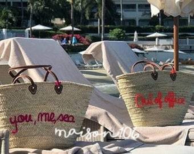 May include: Two large straw tote bags with leather handles. One bag has the text "you, me, sea" embroidered in pink thread. The other bag has the text "Oui, oui, oui" embroidered in pink thread. Both bags are sitting on white beach chairs.