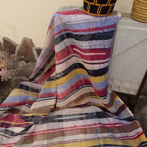 May include: A striped woven throw blanket in shades of red, yellow, blue, black, and white. The blanket is draped over a stool with a black and yellow woven basket on top.