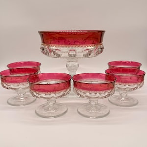 Puede incluir: Un juego de cuencos de postre de cristal transparente y un cuenco con pedestal a juego, cada uno con una banda roja oscura en el borde superior y un patrón texturizado de puntos circulares en relieve en la base, sobre tallos de cristal.